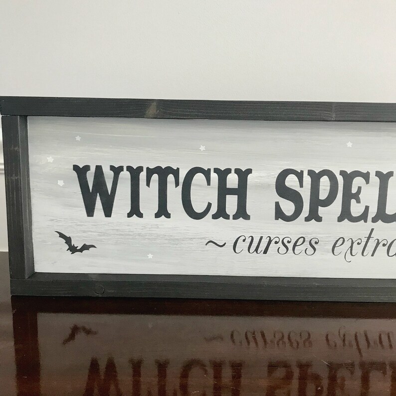 Puede incluir: Un letrero de madera enmarcado en negro con fondo blanco y letras negras que dice "WITCH SPELLS ~ curses extra". El letrero tiene una peque&ntilde;a silueta de murci&eacute;lago negro y estrellas blancas pintadas.