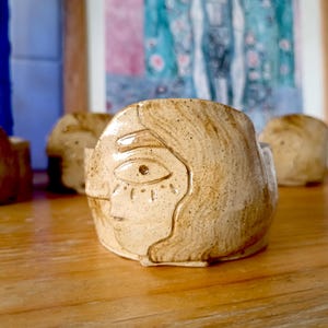 May include: A ceramic planter shaped like a face with a single eye and a textured surface. The planter is beige with brown speckles.