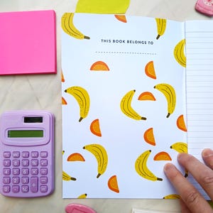 May include: A white notebook with a banana and orange slice pattern. The cover has the text "This book belongs to" printed on it. The notebook is open to a blank page with lined paper.
