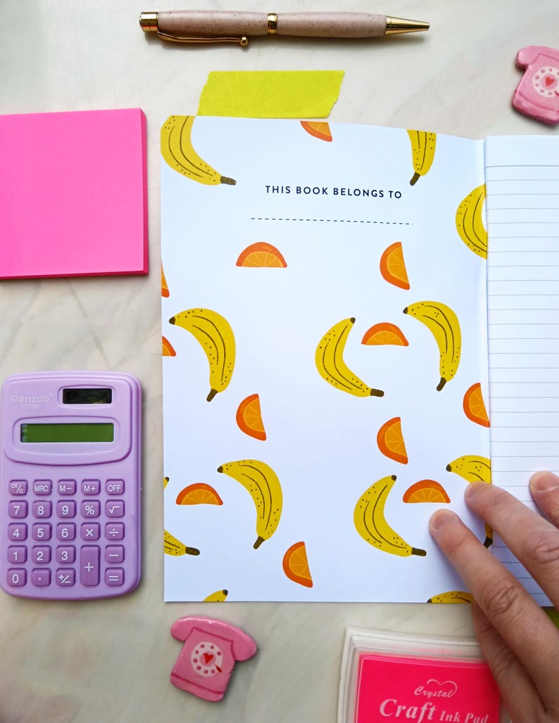 May include: A white notebook with a banana and orange slice pattern. The cover has the text "This book belongs to" printed on it. The notebook is open to a blank page with lined paper.