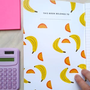 May include: A white notebook with a banana and orange slice pattern. The cover has the text "THIS BOOK BELONGS TO" with a line for writing a name.