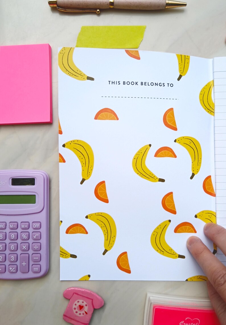 May include: A white notebook with a banana and orange slice pattern. The cover has the text "THIS BOOK BELONGS TO" with a line for writing a name.