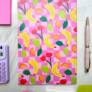 May include: A pink and white checkered notebook with a repeating pattern of yellow bananas, green leaves, red cherries, and orange citrus slices. The notebook has a pink cover and is perfect for taking notes or journaling.