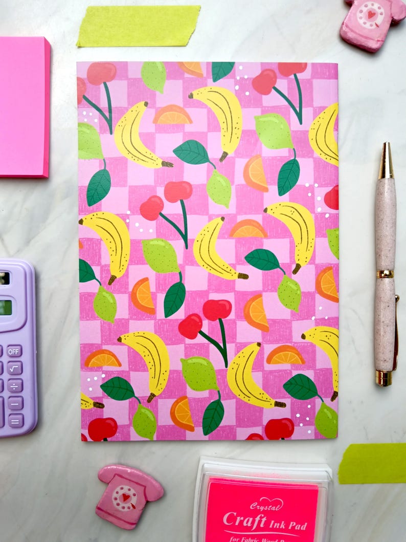 May include: A pink and white checkered notebook with a repeating pattern of yellow bananas, green leaves, red cherries, and orange citrus slices. The notebook has a pink cover and is perfect for taking notes or journaling.