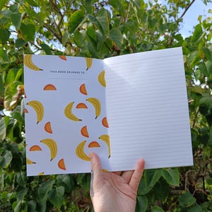 May include: A white notebook with a yellow banana and orange slice pattern. The front cover has the text "This book belongs to".