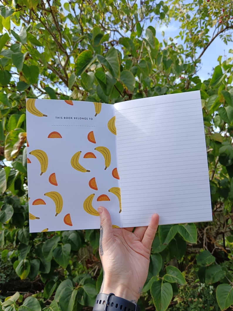 May include: A white notebook with a yellow banana and orange slice pattern. The front cover has the text "This book belongs to".
