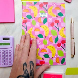 May include: A pink notebook with a checkered pattern and a repeating design of yellow bananas, green leaves, and red cherries. The notebook has a sticker that says "Squeeze the Day!" and is lying on a white surface.