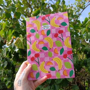 May include: A pink and white checkered notebook with a pattern of yellow bananas, green leaves, orange slices, and red cherries. The notebook has the text "Squeeze the Day!" in pink letters.