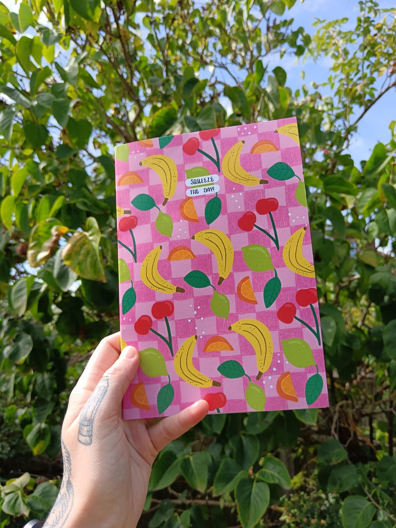 May include: A pink and white checkered notebook with a pattern of yellow bananas, green leaves, orange slices, and red cherries. The notebook has the text "Squeeze the Day!" in pink letters.