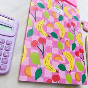 May include: A pink notebook with a checkered pattern featuring bananas, lemons, and cherries. The notebook has a sticker that says "SQUASH THE DRAMA".