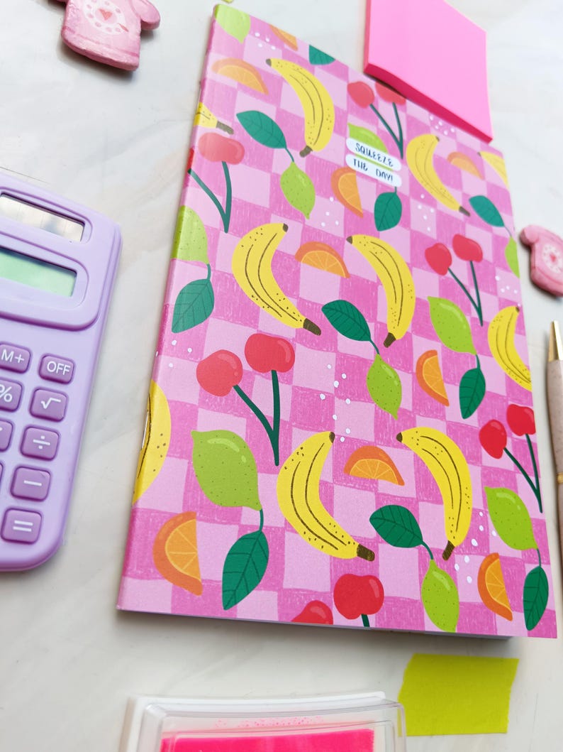May include: A pink notebook with a checkered pattern featuring bananas, lemons, and cherries. The notebook has a sticker that says "SQUASH THE DRAMA".