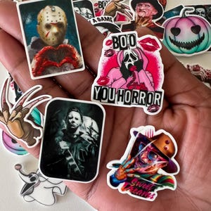 Pode incluir: Uma coleção de autocolantes com temática de terror apresentando personagens icónicas como Jason Voorhees, Michael Myers e Freddy Krueger, ao lado de um autocolante rosa Ghostface com o texto BOO YOU HORROR e Sweet Dreams.