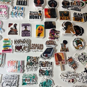 May include: A collection of colorful stickers with various designs and text. The stickers feature images of Black women, inspirational quotes, and phrases like "Mama," "Blessed," and "Black Lives Matter." The stickers are arranged on a white surface.