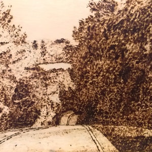 May include: A woodburned image of a winding road leading through a forest with a lake in the distance.