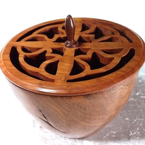 May include: A wooden box with a carved lid featuring a geometric design. The box is made of light brown wood and has a dark brown knob on the top.