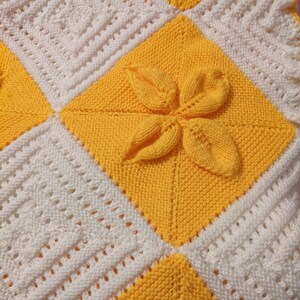 May include: A knitted baby blanket with a yellow and white pattern. The blanket is made of squares with a flower design in the center of each square.