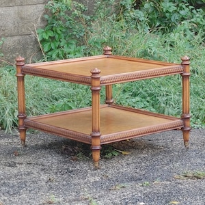 May include: A two-tiered, square wooden side table with ornate detailing. The table has a light brown finish, with carved accents around the edges of the shelves and on the legs. The table is approximately 24 inches wide and 24 inches long.