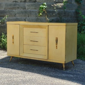 May include: A light wood sideboard with three drawers and two doors. The doors have gold handles. The sideboard has four gold legs.