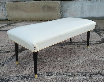 May include: A white upholstered bench with black legs and gold accents. The bench is sitting on a gray concrete surface.