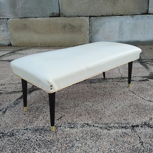 May include: A white upholstered bench with black legs and gold accents. The bench is sitting on a gray concrete surface.