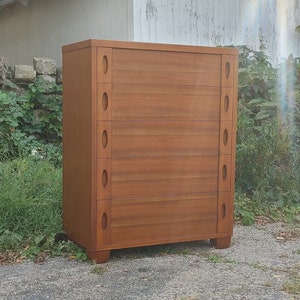 May include: A five-drawer dresser made of wood with a light brown finish. The dresser has oval-shaped drawer pulls and a simple design.