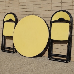 May include: A set of three yellow folding chairs and a round yellow table with black metal frames. The chairs and table are all in a folded position.