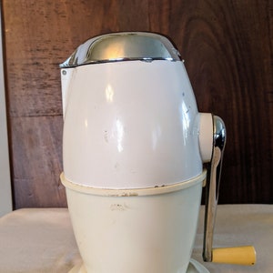 May include: A white manual ice crusher with a chrome top and a yellow handle. The crusher has a base and a cylindrical body with a crank on the side.