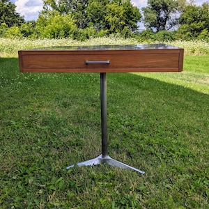 May include: A wooden end table with a single drawer and a silver metal base. The table is standing on a grassy lawn.