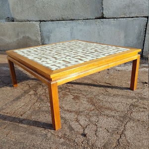 May include: A square coffee table with a wood frame and a tiled top. The tiles are a light beige color with brown accents. The table is sitting on a gravel surface.