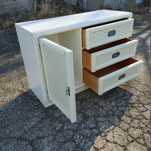 May include: White wicker cabinet with three drawers and a door. The door is open, revealing a shelf inside. The drawers have metal handles.
