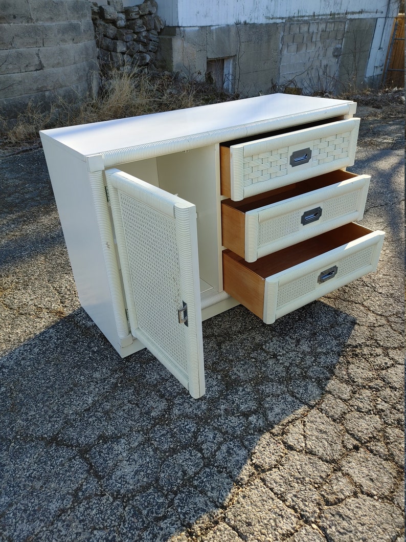 May include: White wicker cabinet with three drawers and a door. The door is open, revealing a shelf inside. The drawers have metal handles.
