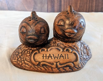 Könnte beinhalten: Ein braunes Keramik-Salz- und Pfefferstreuer-Set in Form von Fischen mit geschlossenen Augen und einem kleinen Mund. Die Streuer sitzen auf einem braunen Sockel mit floralem Design und dem Wort "HAWAII" in einer dekorativen Schrift.