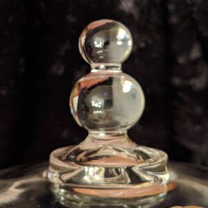 Puede incluir: Un tap&oacute;n de botella de vidrio transparente con una parte superior redondeada y una base con un dise&ntilde;o decorativo.