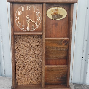 May include: A wooden wall organizer with a clock, corkboard, and three compartments. The clock face is white with black numbers and hands. The corkboard is brown and has a rough texture. The compartments are made of wood and have a natural finish.