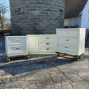 May include: A set of three white wicker dressers with brass hardware. The dressers are on wheels and have a distressed finish. The set includes a large dresser, a medium dresser, and a small nightstand.