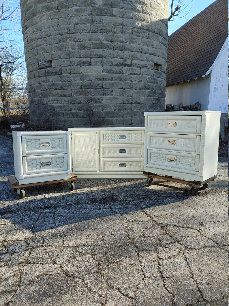 May include: A set of three white wicker dressers with brass hardware. The dressers are on wheels and have a distressed finish. The set includes a large dresser, a medium dresser, and a small nightstand.