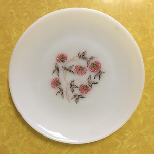 May include: White ceramic plate with a floral design. The plate features a central pattern of pink flowers with green leaves and brown stems. The plate is round and sits on a yellow surface.