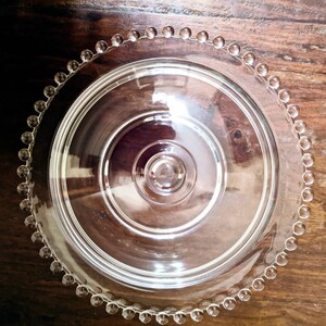 Puede incluir: Un soporte para tartas de vidrio transparente con un borde de cuentas. El soporte tiene una tapa abovedada y un pedestal central.