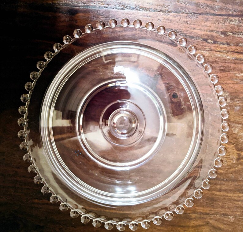 Puede incluir: Un soporte para tartas de vidrio transparente con un borde de cuentas. El soporte tiene una tapa abovedada y un pedestal central.