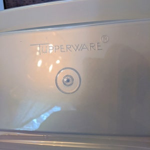 Op de afbeelding: Een witte plastic container met het woord "Tupperware" erop gedrukt. De container heeft een doorzichtig plastic deksel met een kleine ronde knop aan de bovenkant.