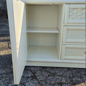 May include: White wicker dresser with a single door open revealing two shelves. The dresser has a woven pattern on the front and sides.