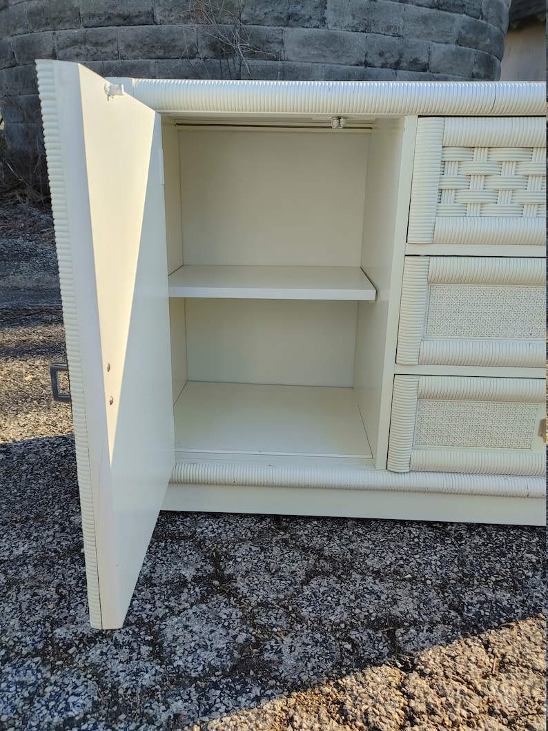 May include: White wicker dresser with a single door open revealing two shelves. The dresser has a woven pattern on the front and sides.