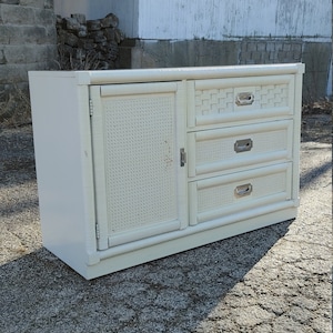 May include: White wicker dresser with three drawers and a cabinet door. The dresser has silver hardware and a woven pattern.