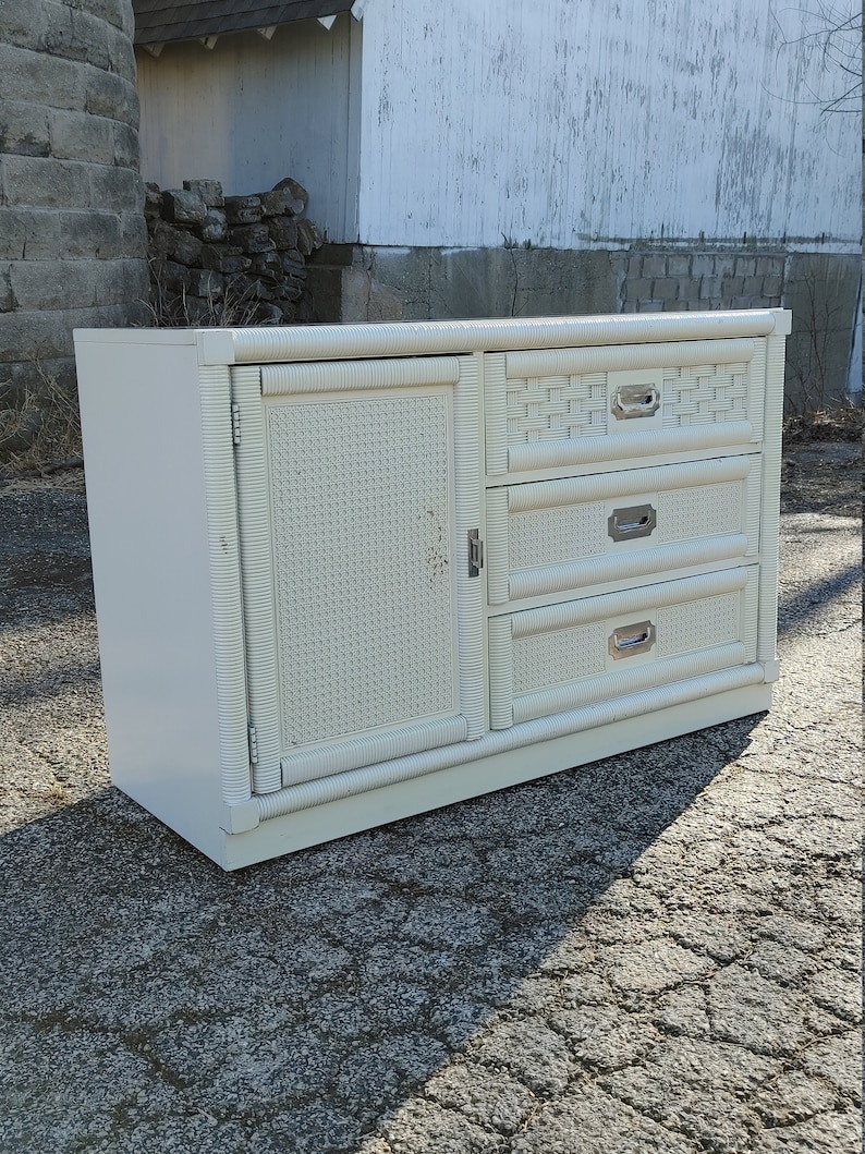 May include: White wicker dresser with three drawers and a cabinet door. The dresser has silver hardware and a woven pattern.