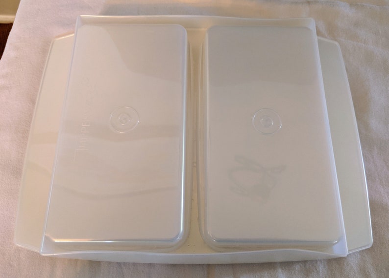 Op de afbeelding: Twee transparante plastic containers met deksels, genest in een witte plastic bak. De containers hebben een cirkelvormig ontwerp aan de bovenkant.