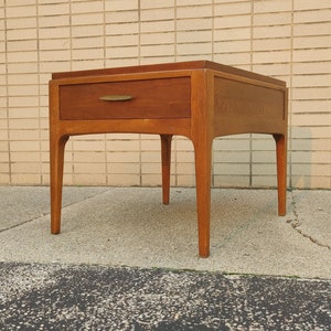 May include: A mid-century modern end table with a single drawer and tapered legs. The table is made of wood and has a light brown finish.