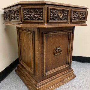 May include: A dark wood octagonal end table with a single door and two drawers. The table has intricate carvings on the front and sides. The drawers have brass knobs.