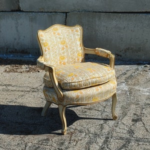 May include: A vintage armchair with a floral patterned upholstery in shades of yellow and white. The chair has a carved wooden frame with a light wood finish.