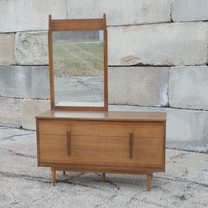 May include: A mid-century modern dresser with a mirror. The dresser is made of wood and has six drawers. The mirror is attached to the top of the dresser and has a simple frame.