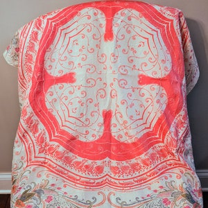 May include: A white tablecloth with a pink and white floral pattern. The tablecloth has a large, central design with a repeating pattern of swirls and leaves. The tablecloth is folded and draped over a chair.